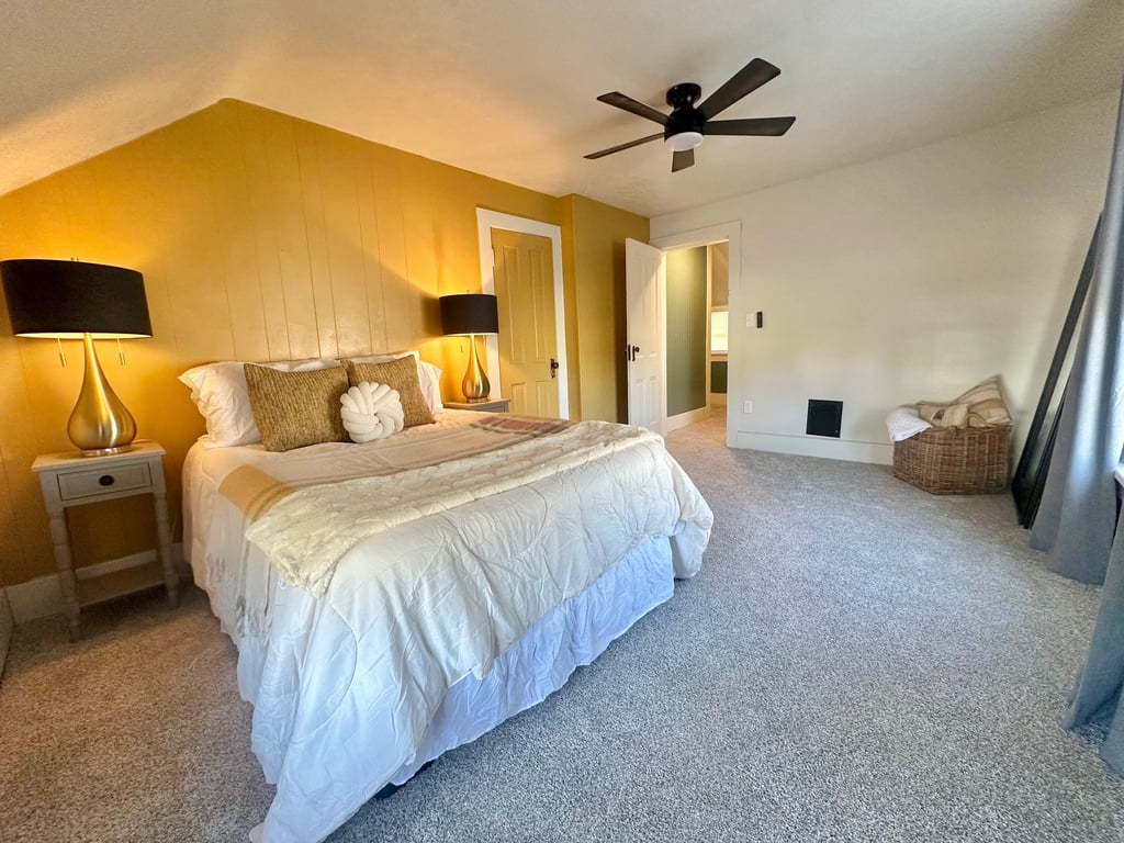 Cozy bedroom with a yellow accent wall, white bedding, gold lamps, and a black ceiling fan.