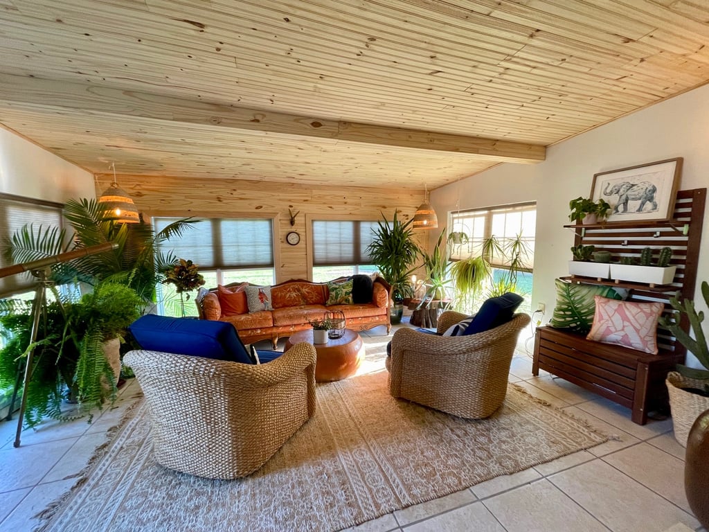 Boho sunroom interior with wood ceiling, wicker chairs, orange sofa, and many indoor plants.
