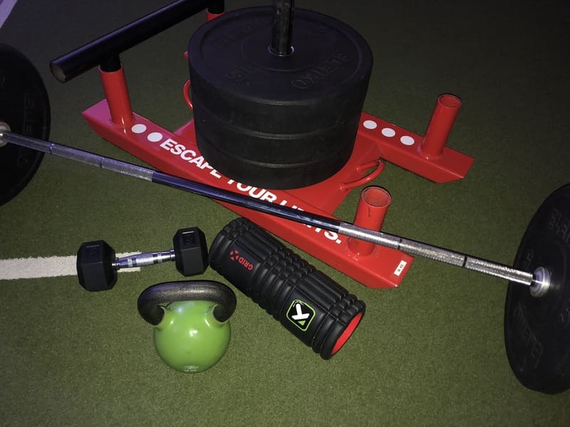 Fitnessgeräte auf Kunstrasen: roter Gewichtsschlitten, Langhantel, grüne Kettlebell und Faszienrolle