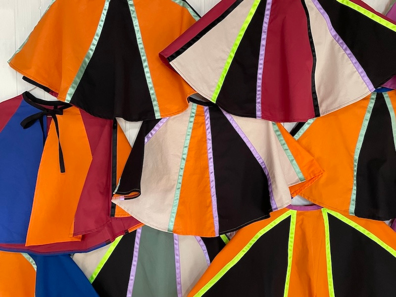 Handmade colorful circle capes with vibrant ribbon stripes in orange, black, and maroon fabric.