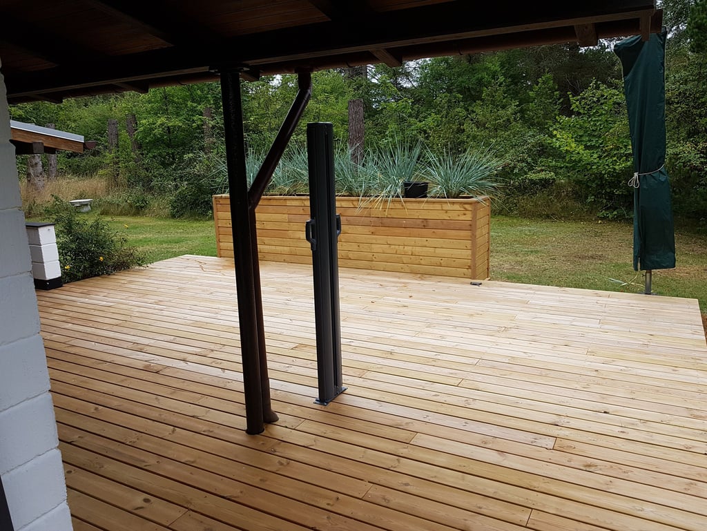 En moderne udendørs træterrasse med en stor plantekasse og en frodig grøn skov i baggrunden.