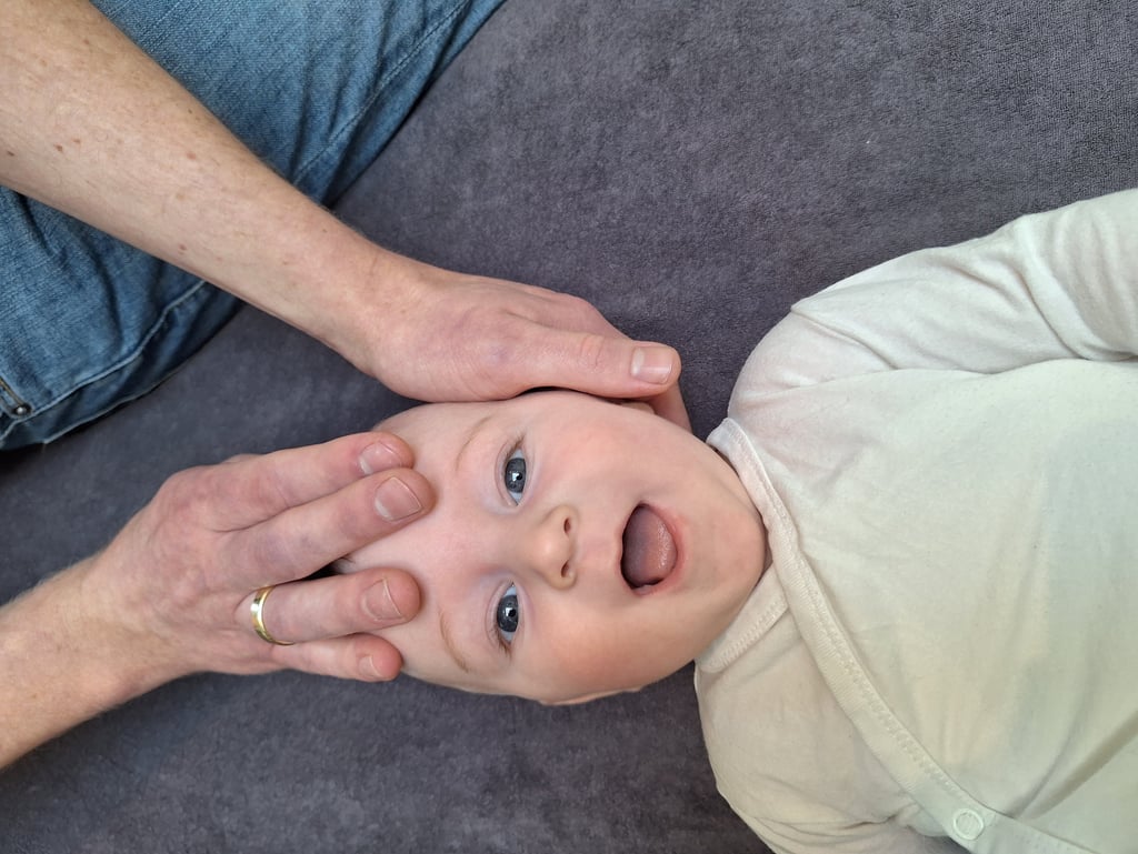 een baby ligt op zijn rug en lacht terwijl 2handen zijn hoofd vasthouden.