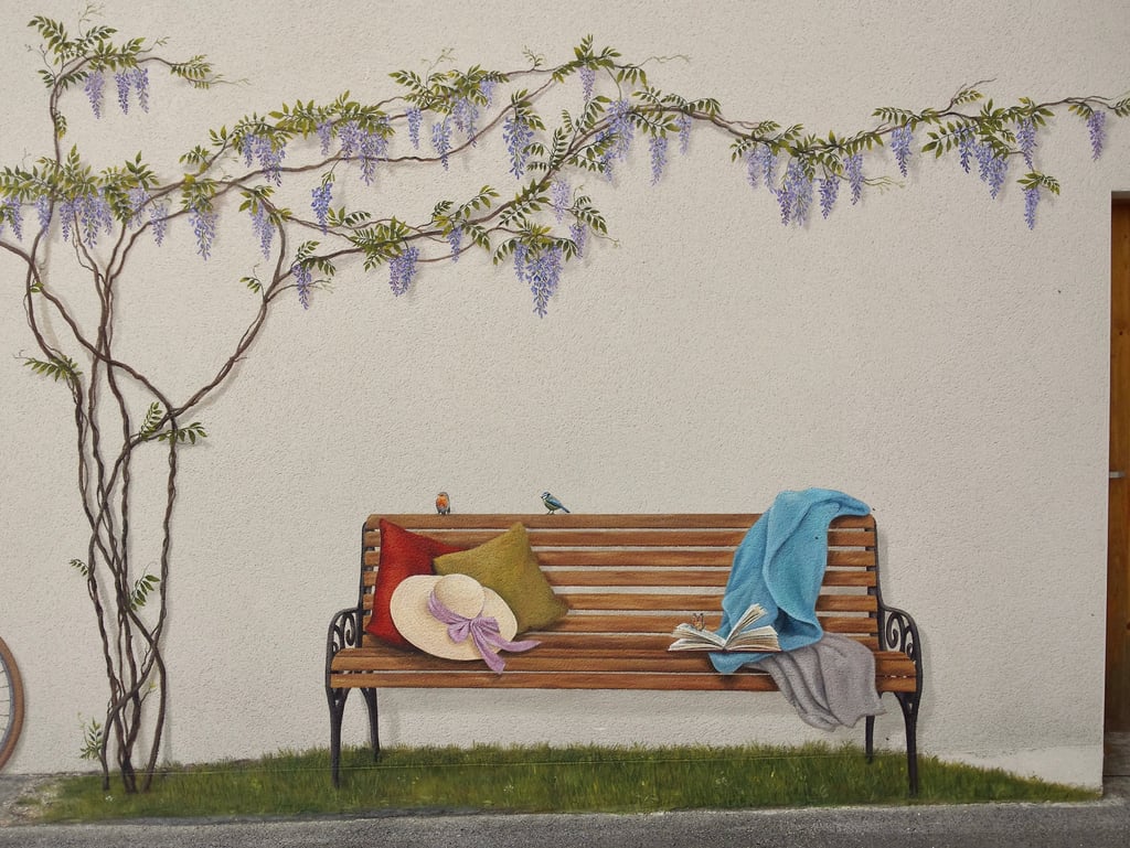 Fresque en trompe-l'oeil sur façade. Un banc posé sur l'herbe, des oiseaux, un livre et une glycine.