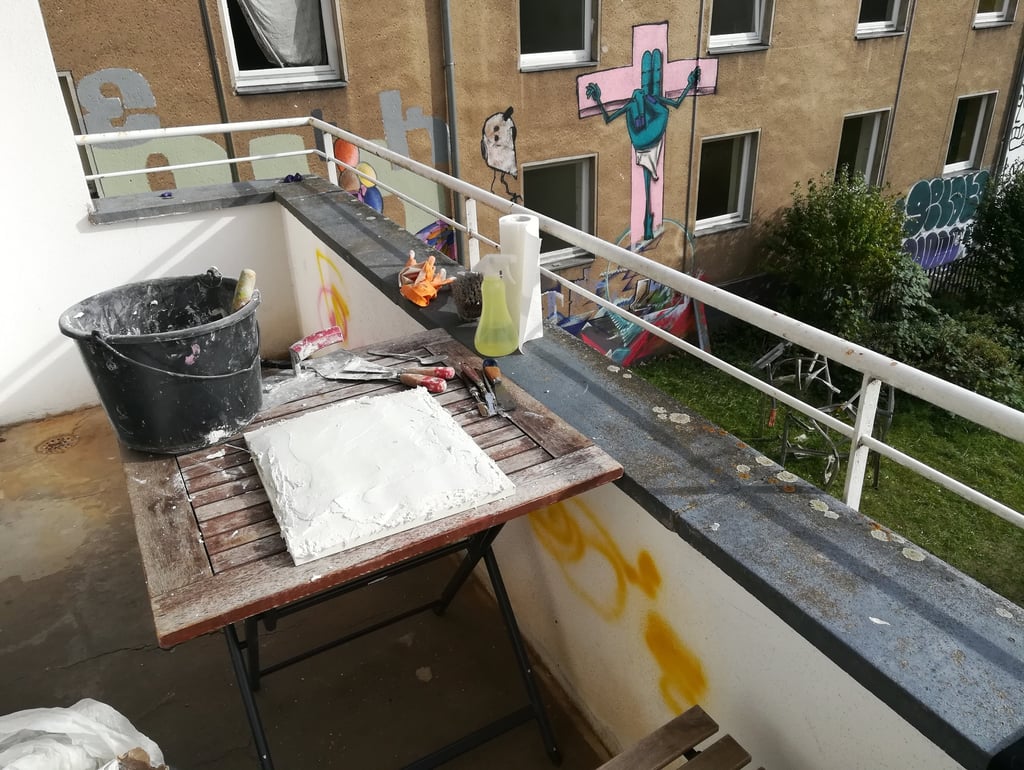 Artist sculpting white plaster on a balcony table overlooking graffiti-covered urban buildings.