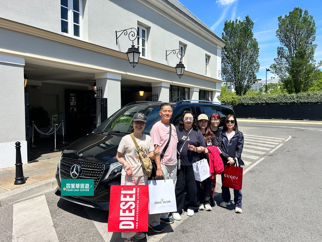 A group of tourists holding luxury brand shopping bags poses next to a black Mercedes-Benz chauffeur van.