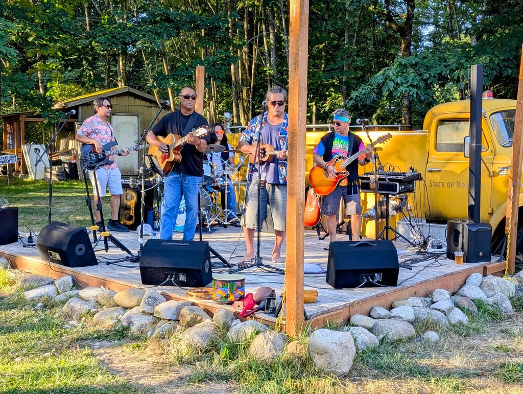 Live band performing outdoor music on a wooden stage with a yellow vintage truck background.