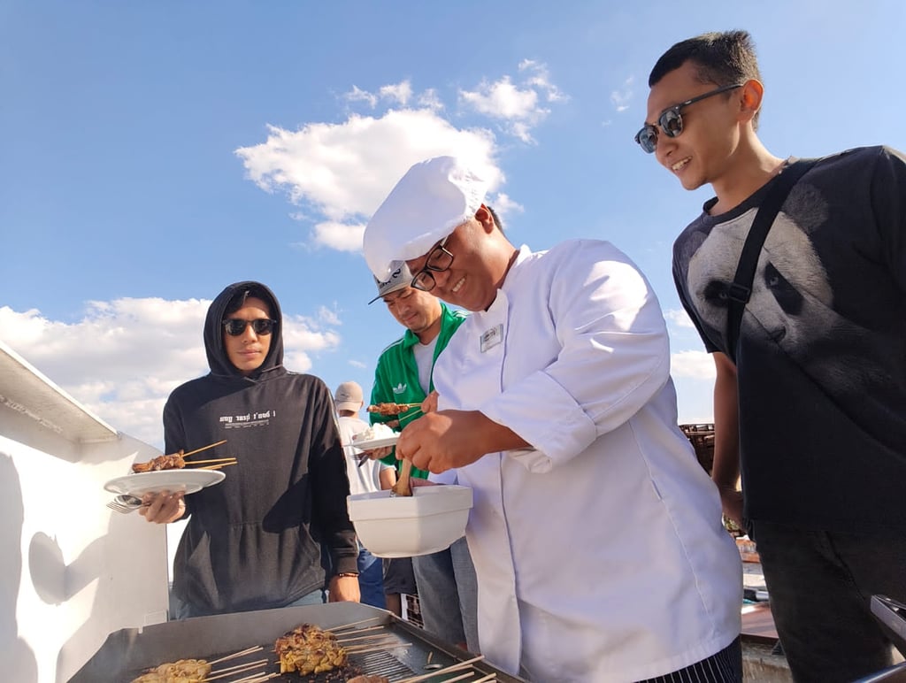 A kitchen crew grills chicken satay skewers for river crew party on a sunny outdoor deck.