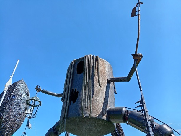 A large rustic metal sculpture of a mechanical character with a lantern and staff against a clear blue sky.