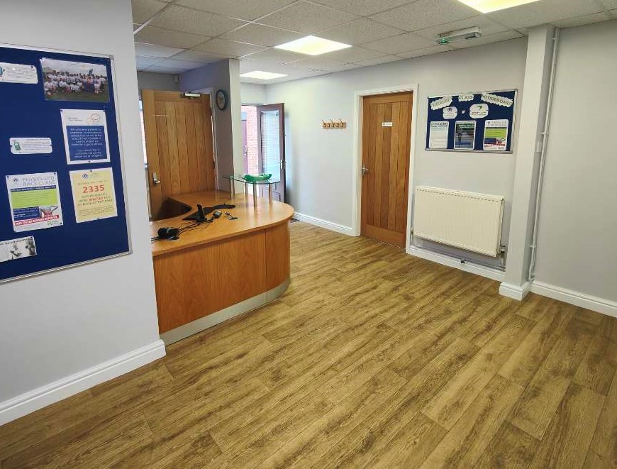 A reception with curved wood desk, wooden flooring/door, and light-grey walls.