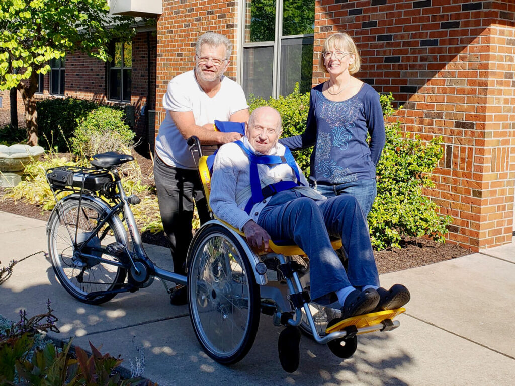 One person sitting in the Giving Rides bike with two people standing behind them smiling