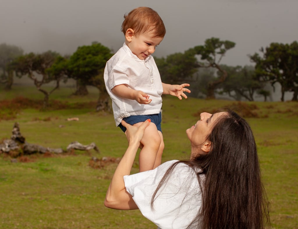 Mother lifting smiling toddler up, green grass, foggy forest and laurel trees in background, joy and play.