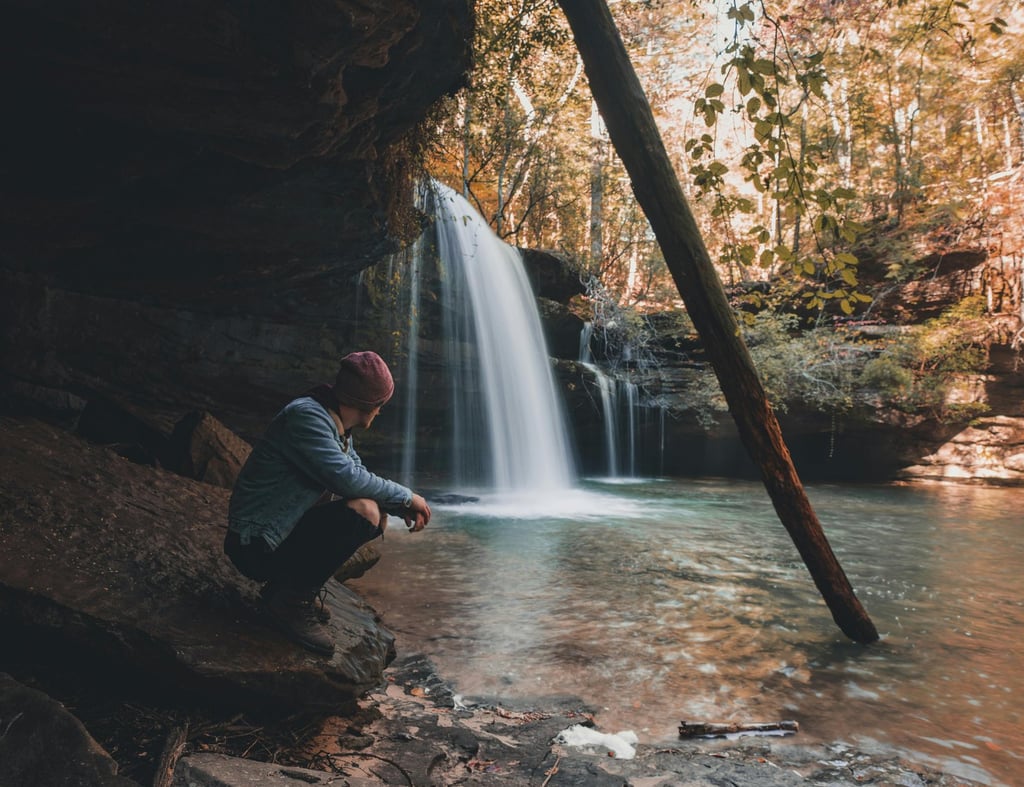 Person resting by waterfall with backpack, showing natural shelter and wilderness hydration