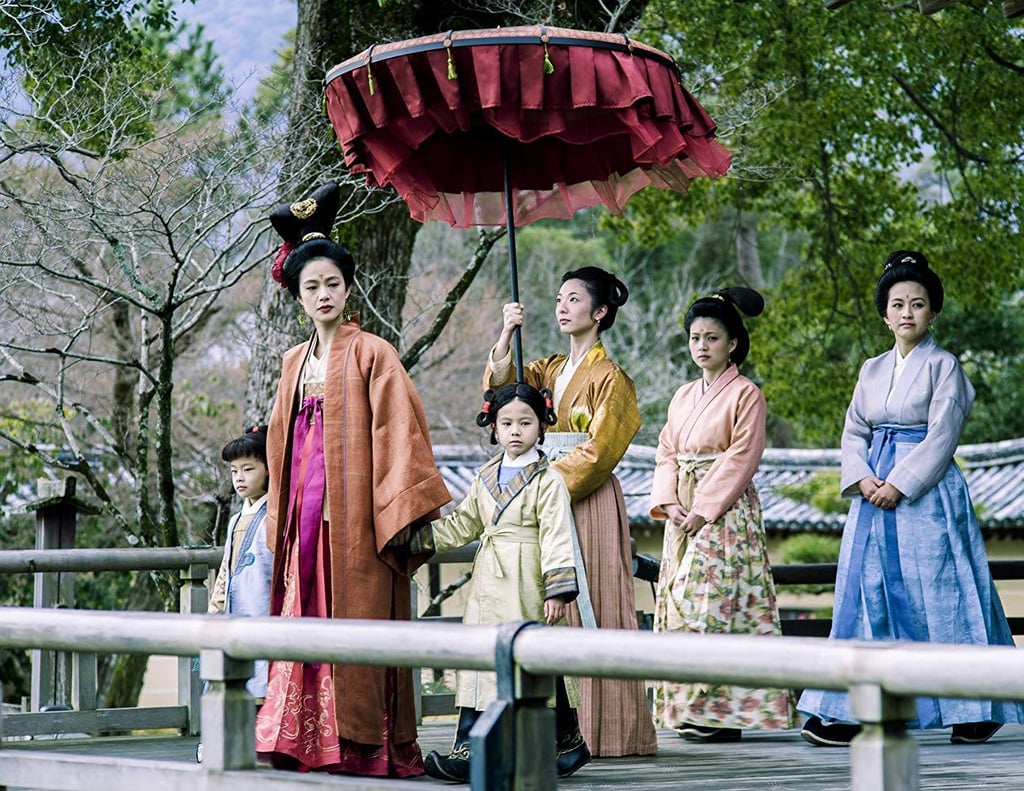Women in traditional Japanese hanfu and kimono walk on a wooden bridge under a red ceremonial umbrella.