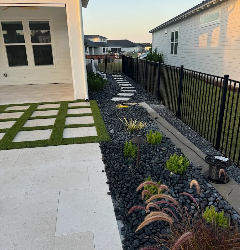 Modern backyard landscaping featuring a travertine patio, black river rock, and artificial turf strips.