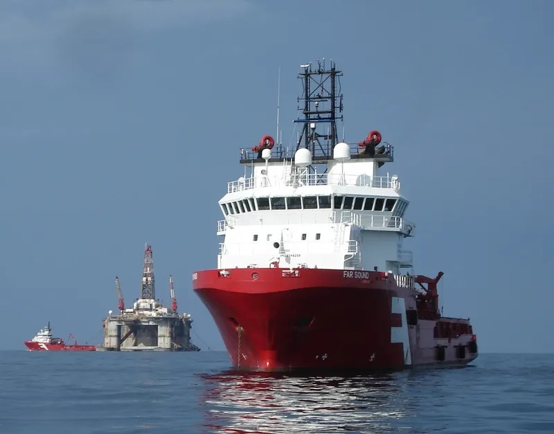Red offshore supply ship Far Sound anchored near a semi-submersible oil drilling rig at sea.