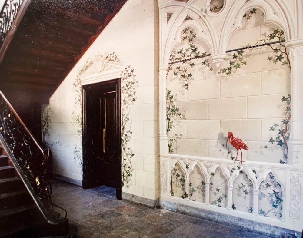 Hallway with wooden staircase and large mural of faux sandstone, ivy, gothic arches, and red bird.