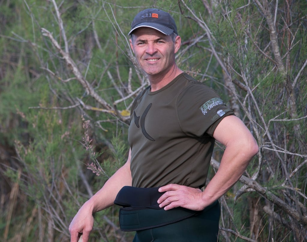 A smiling male outdoorsman in a green t-shirt and 5.11 cap standing in nature.
