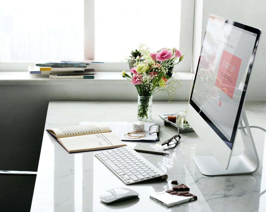 A bright workspace with keyboard, monitor, mouse, desk accessories, and flowers in a vase next to a window.