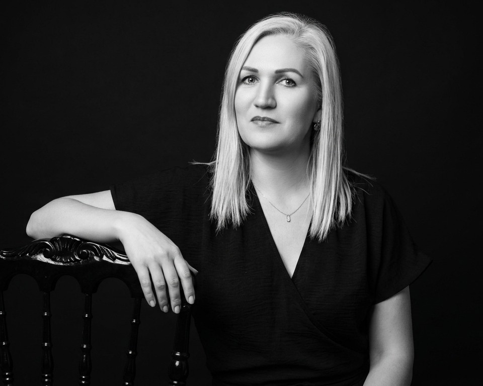 Black and white professional headshot of a blonde woman leaning on a chair against a dark background.