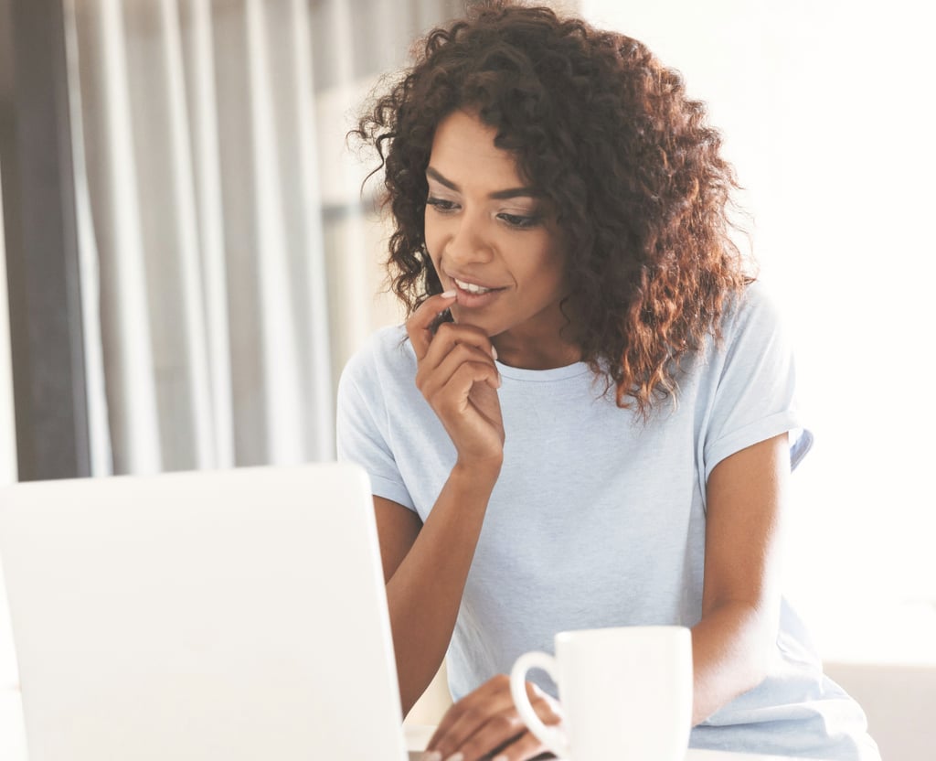 A smiling African American woman with curly hair working on a laptop at home with a coffee mug.