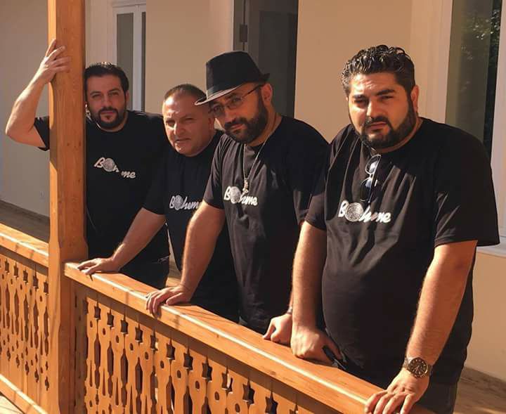 Four men in matching black Bohème t-shirts posing on a wooden balcony railing outdoors.