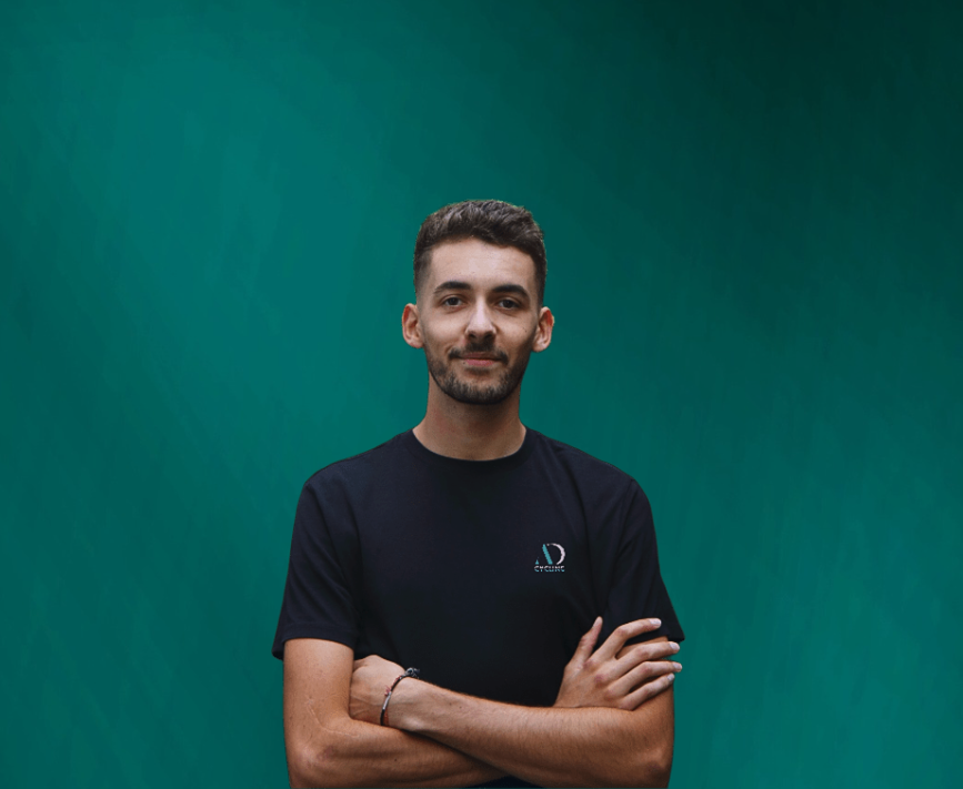 A young man in a black AD Cycling t-shirt posing with crossed arms against a teal background.
