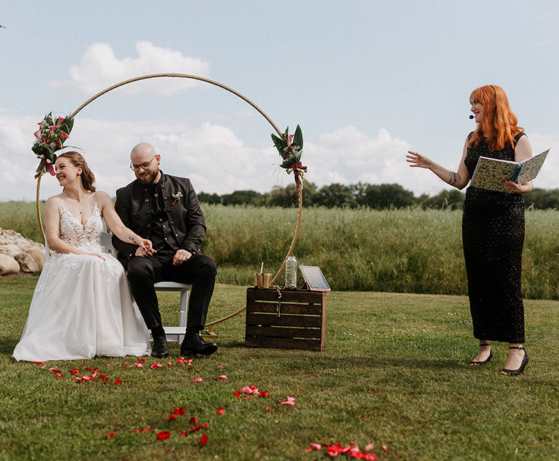 grinsend sitzen Braut und Bräutigam nebeneinander während die Traurednerin im schwarzen Glitzerkleid eine Anekdote erzählt
