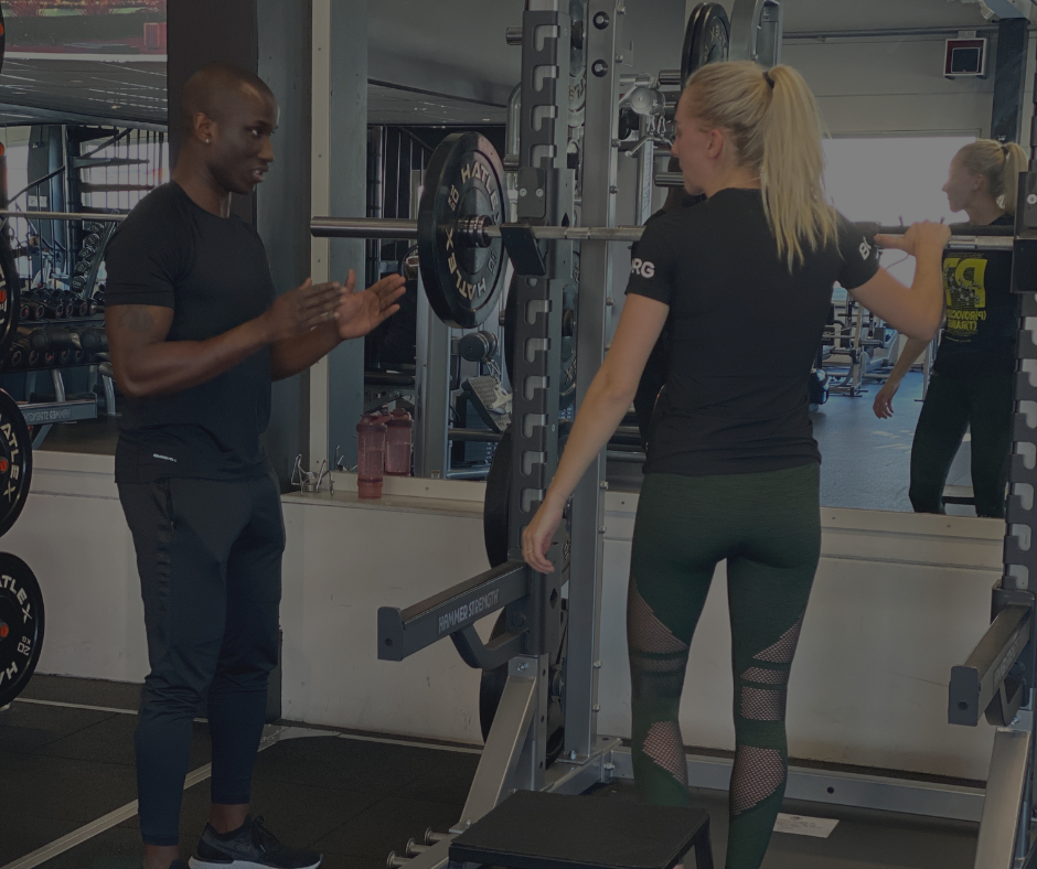 A personal trainer providing coaching to a woman performing barbell squats at a gym power rack.
