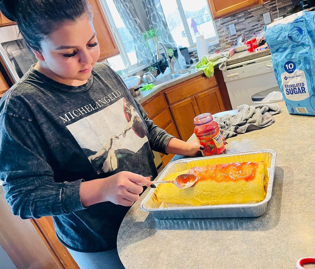 A woman spreads strawberry jam over a rolled sponge cake in a kitchen to make a Swiss roll dessert.