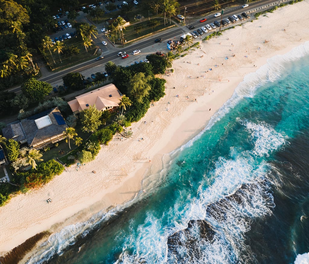 Aerial view of a tropical white sand beach with turquoise ocean waves crashing near coastal houses and a road.