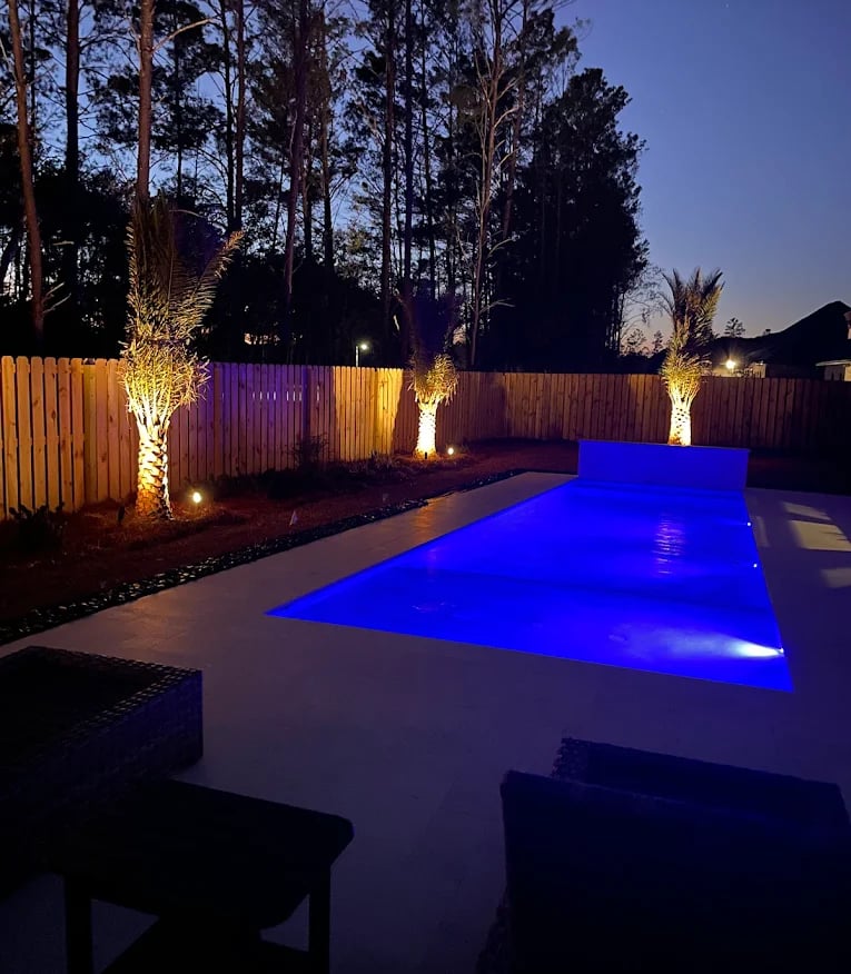 Modern backyard swimming pool with vibrant blue LED lighting and illuminated palm trees at night.