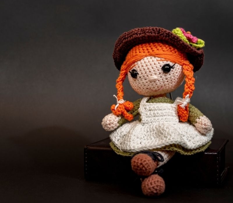 Muñeca tejida a crochet hecha a mano con trenzas rojas y vestido vintage sentada sobre una caja