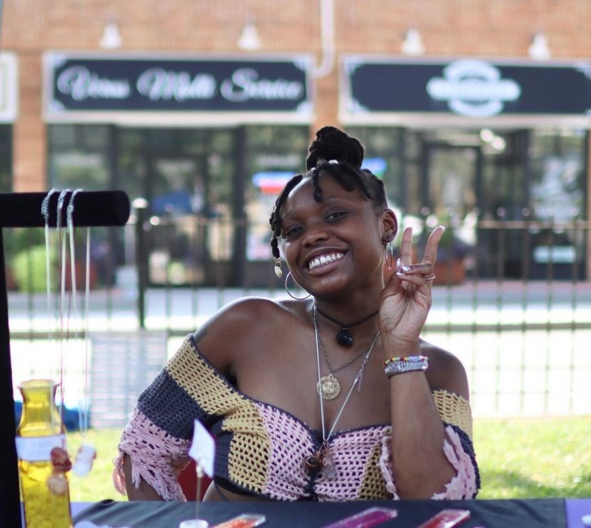 Smiling woman entrepreneur at outdoor craft market table with handmade jewelry and crochet top.