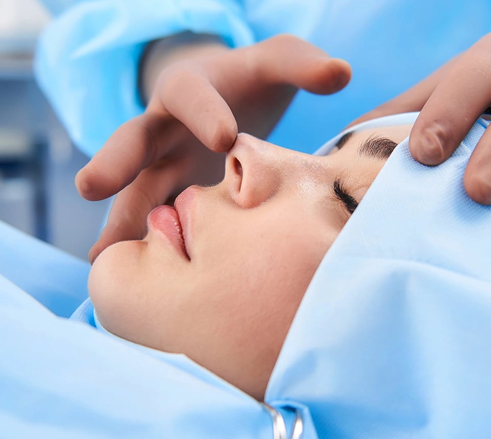 Patient draped in blue, surgeon's hands examine nose for rhinoplasty.