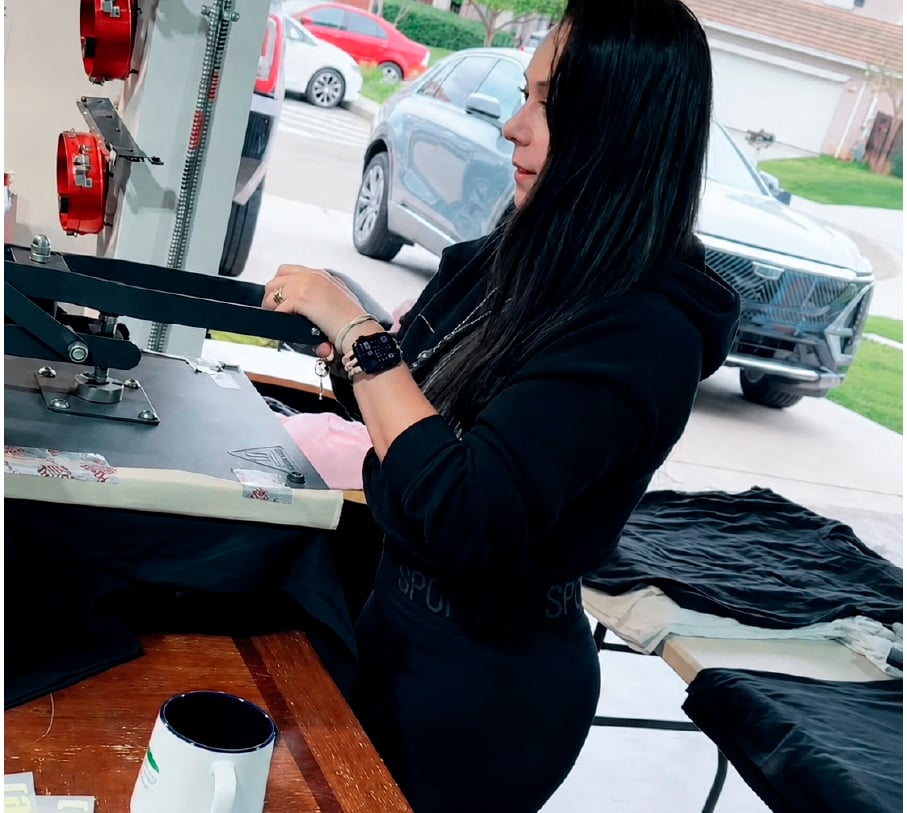 A woman uses a professional heat press machine to print custom designs onto black t-shirts.