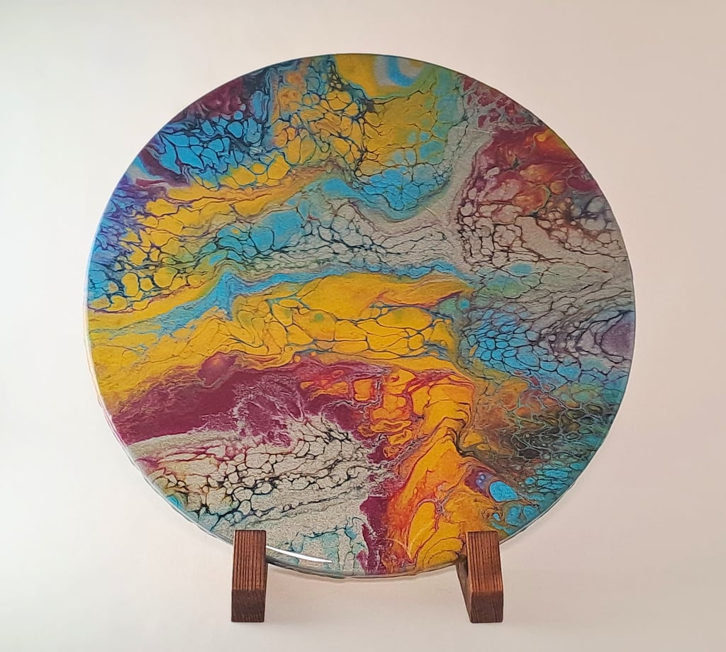 Circular acrylic pour painting with vibrant yellow, blue, and red cells on a wooden display stand.
