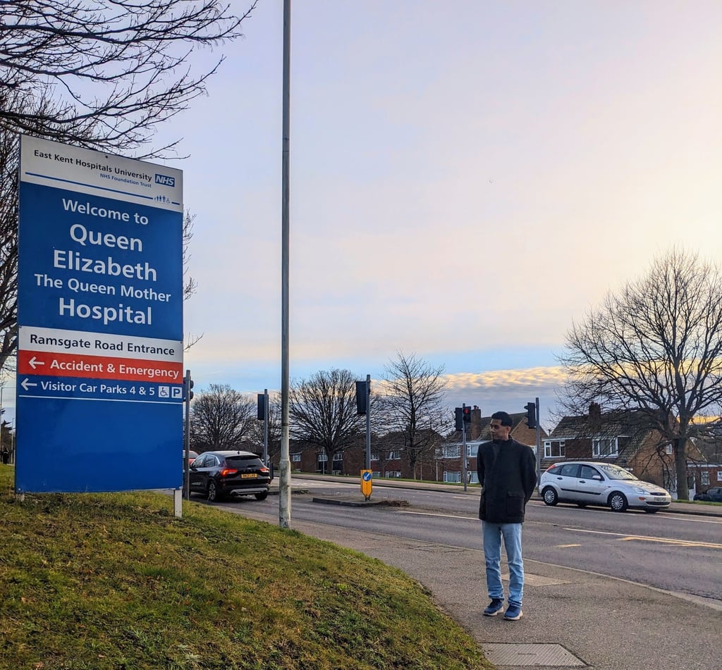 A man standing at a distance looking at the signpost reading 'Welcome to Queen Elizabeth The Queen Mother Hospital'