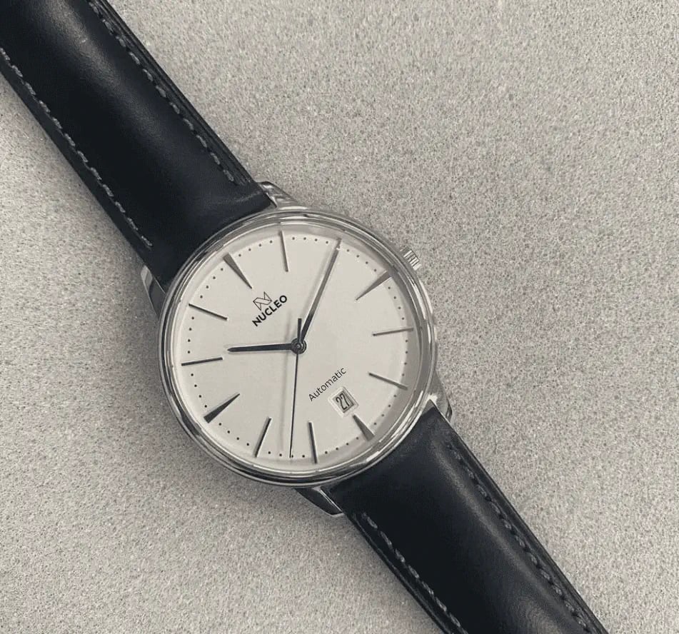 orologio dress nucleo con quadrante bianco indici e lancette argento data a ore 6 cinturino pelle nera posto su piano grigio