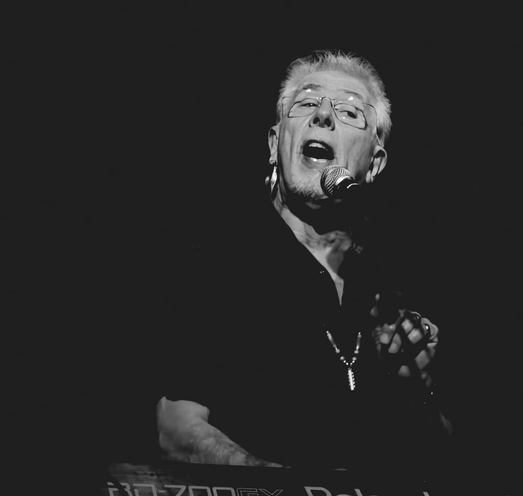 Black and white portrait of a male musician singing into a microphone while playing a keyboard.