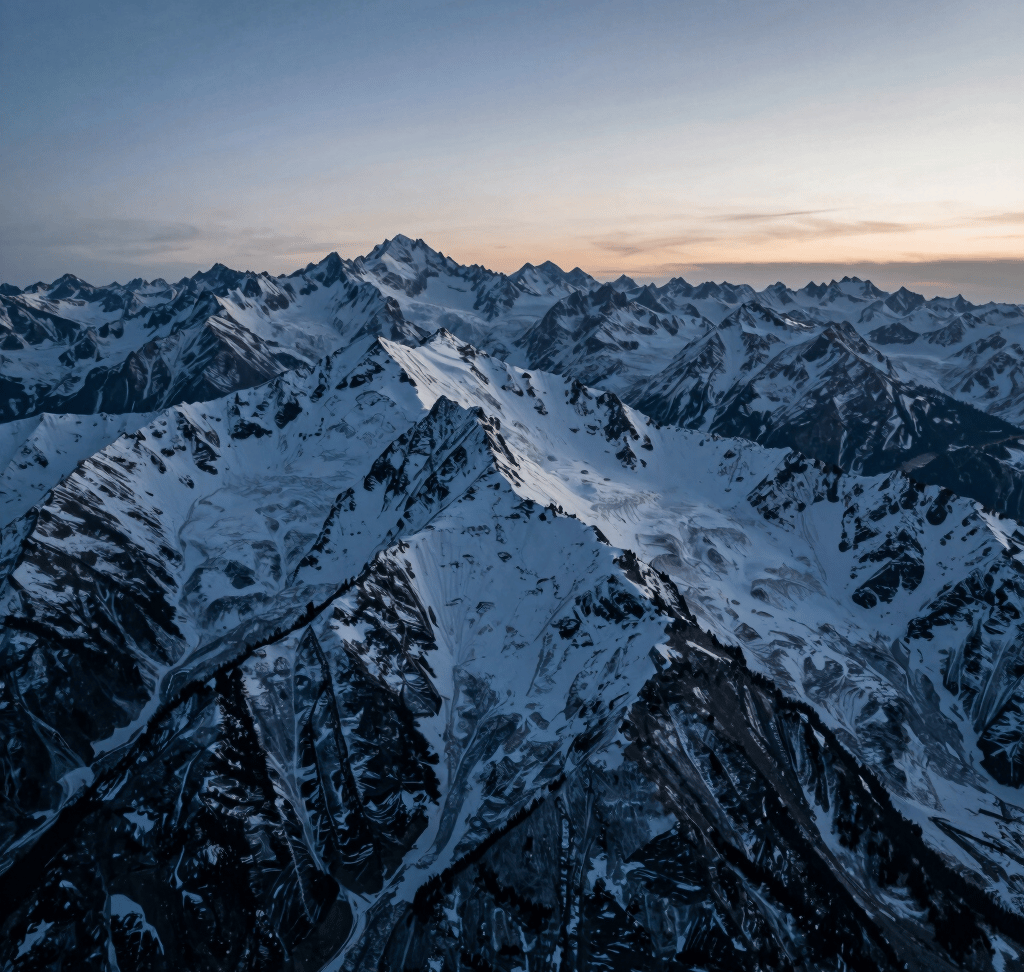 A high-altitude cinematic drone shot of a rugged mountain ridge at blue hour. The lighting transitions from deep #1B263B shadows to soft #A0B0C0 highlights on the snow-capped peaks. The composition is wide and sweeping, capturing professional 4K clarity and a sense of epic scale.