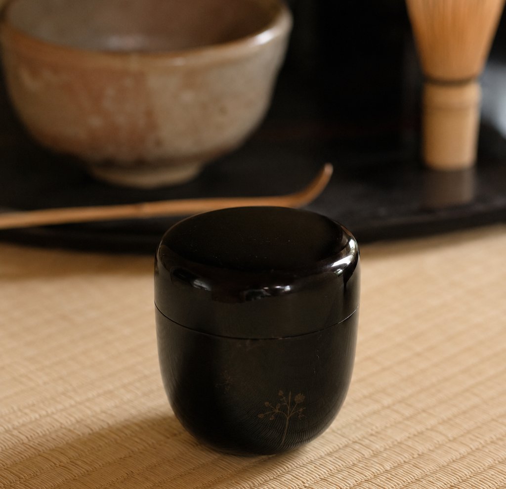 A black lacquer natsume tea caddy on a tatami mat with a matcha bowl and bamboo whisk.