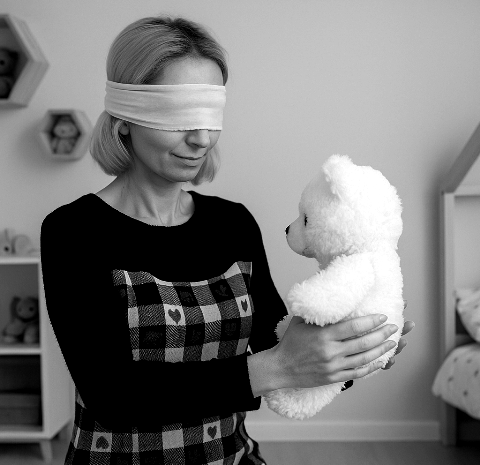 Kobieta z opaską na oczach stoi w jasnym, skromnym pokoju dziecięcym i delikatnie trzyma w dłoniach