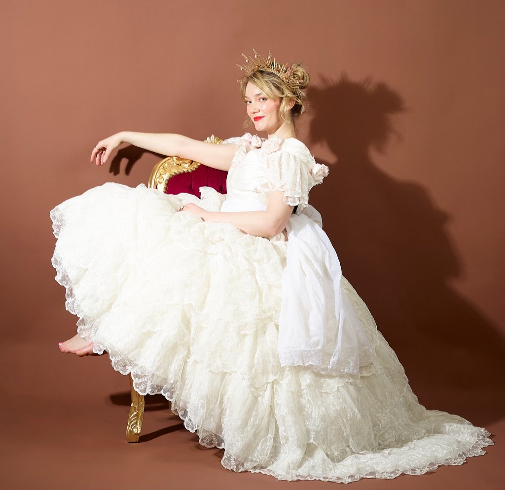 A woman in a ruffled white ballgown and gold crown posing on a vintage velvet chair.
