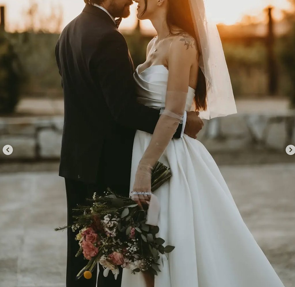 Novios abrazados al atardecer con ramo en tonos rosados.