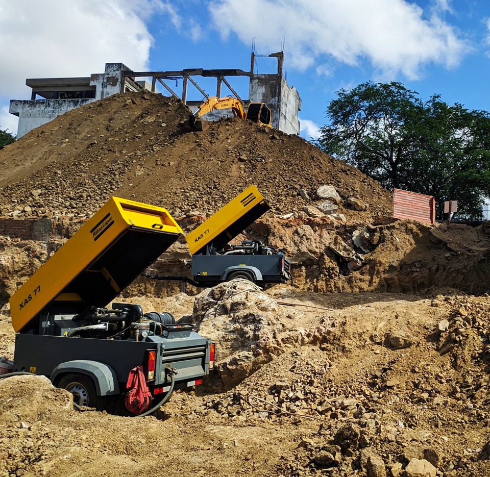 Dois compressores amarelos em uma obra de terraplanagem, realizando serviço de desmonte de rochas