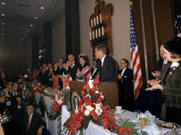 JFK in a suit and tie standing at a podium with a microphone