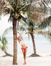 A blonde model wearing a white crop top, red bikini bottoms, and luxury jewelry poses against beach