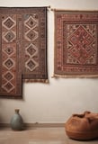 Colorful woven rugs with intricate geometric patterns hang vertically on a wooden rail against a rustic stone and plaster wall. A window with green shutters is partially visible above, and cobbled stones lie at the base.
