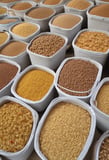 A series of transparent dispensers filled with various types of grains and seeds, each labeled with its name and price per kilogram. The containers include nut-free muesli, muesli base, porridge oats, jumbo oats, wheat flakes, rye flakes, barley flakes, red lentils, sunflower seeds, and whole-wheat penne. The setup is likely located in a shop or market with visible brick walls in the background.