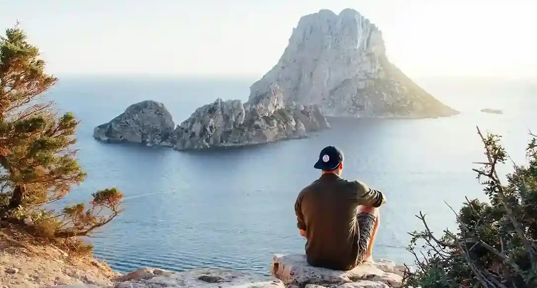 Homme avec 1 casquette visière en AR assis sur 1 rocher face à 1 gros promontoir rocheux dans la mer